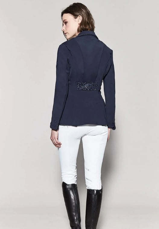 Woman wearing a navy blazer with embellishments, white pants, and black riding boots on a light gray background